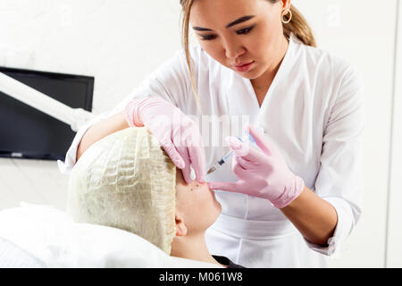Dark-haired woman an asian beautician pushes a syringe to inject Botox into the cheekbones of a young woman to correct the form in the medical office Stock Photo