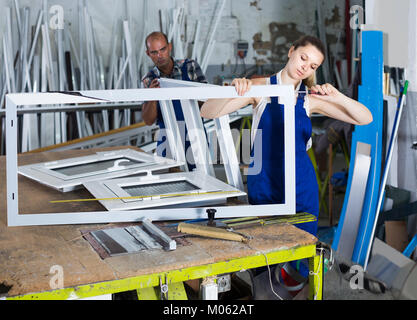 Young female worker assembling metal-plastic window in workshop, using screwdriver Stock Photo