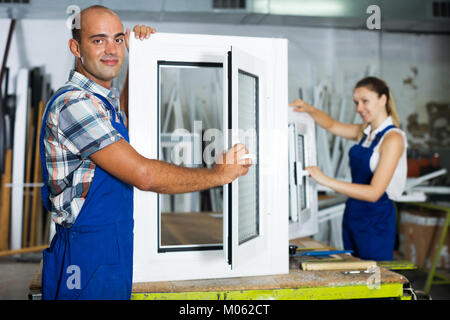 Smiling workman in blue overalls demonstrating pvc window in workshop Stock Photo