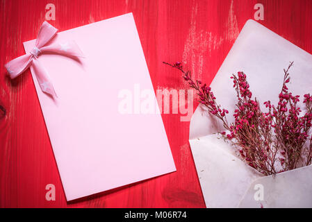 Greeting card idea with an open envelope full of beautiful red flowers near an empty pink paper note with a ribbon bow, on a red wooden background. Stock Photo