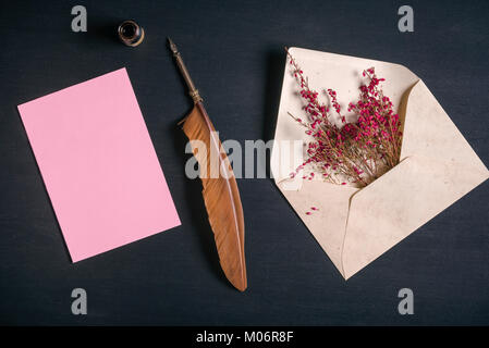 Vintage envelope full with pink flowers, an antique quill pen, an ink pot and a blank message card, on a black wooden background. Stock Photo