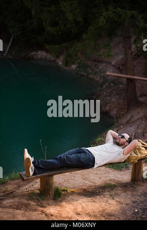 Man Relaxing Outside. Wearing a white T shirt, dark pants, sunglasses, a young guy is lying down on wooden bench against lake in a forest outdoors Stock Photo