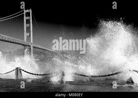 Golden Gate Bridge. Huge waves crash over the sea wall below the bridge in the foreground . Black and white monochrome with dark sky. Copy Space. Stock Photo