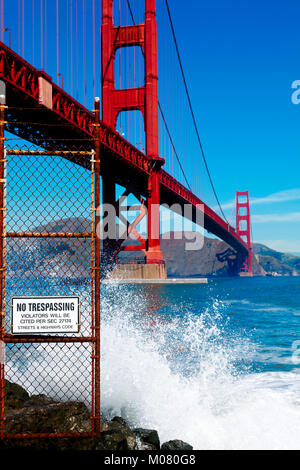 Golden Gate Bridge seen from below. Sign that reads No Trespassing  next to dangerous slippery rocks and surf. Stock Photo