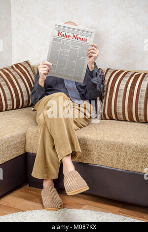 A man wearing glasses is sitting on a couch at home, reading a newspaper reporting fake news. Fake Lorem ipsum text. Stock Photo