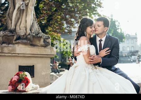 Sensitive portrait of the adorable newlywed couple smiling and tenderly hugging on the old fountain. Stock Photo