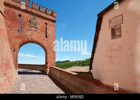 Barolo medieval castle entrance arch in red bricks and emblem with empty street and green hill in a sunny summer day Stock Photo