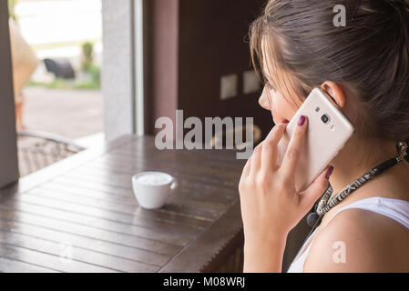 Girl talk to a cell phone in a coffee shop. View behind girl shoulder. Front of is table with a cup of coffee. Stock Photo