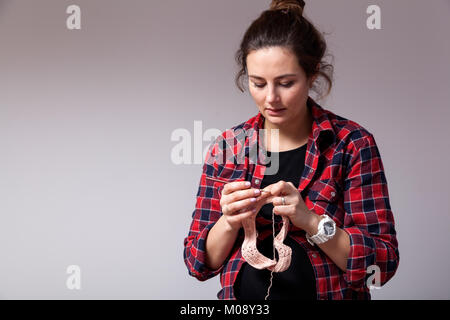 Young dark-haired married woman in plaid shirt knits a pink cap on a gray isolated background Stock Photo