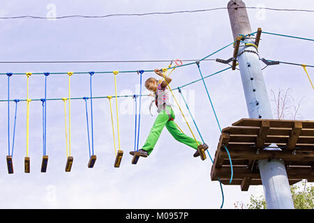 A teenage girl confidently moves on swinging boards at a height in a rope park. Courage and overcome Stock Photo