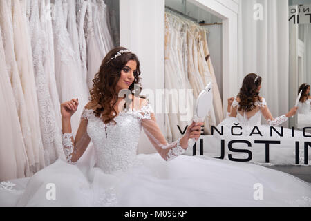 one person, young bride, looking at herself in mirror, bridal salon, wearing gown. Stock Photo