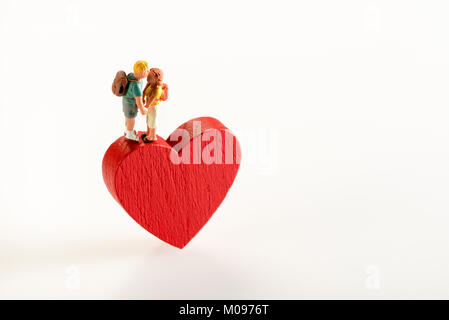Conceptual close-up of a miniature figure couple in love holding hands while standing on a red heart wooden shape against white background for copy sp Stock Photo