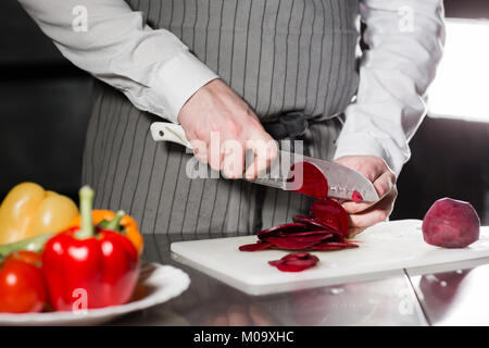 Young chef cutting beet on a white cutting board. Closeup of hand with knife cutting fresh vegetable. Cooking in a restaurant kitchen Stock Photo