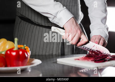 Closeup of hand with knife cutting fresh vegetable. Young chef cutting beet on a white cutting board closeup. Cooking in a restaurant kitchen Stock Photo