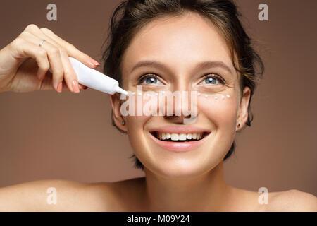 Beautiful smiling woman using cream around the eyes. Photo of woman with perfect skin on brown background. Skin care and beauty Stock Photo