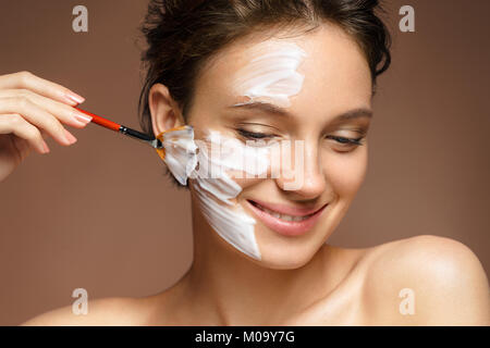 Young woman with moisturizing facial mask and hands of the beautician on brown background. Photo of pretty woman receives the spa treatments. Beauty & Stock Photo