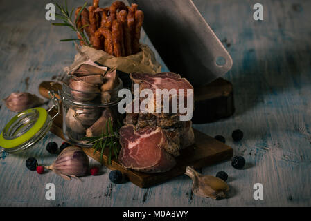 Horizontal photo of stacked pieces of dried smoked meat from italian wild boar. Meat is on vintage chopping board and blue wooden table with sausages, Stock Photo