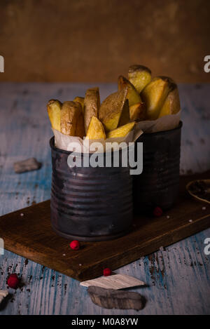 Vertical photo of fried potatoes. Potatoes were roasted with skin. Food is placed in two vintage metal cans which is filled by paper. Cans are on dark Stock Photo