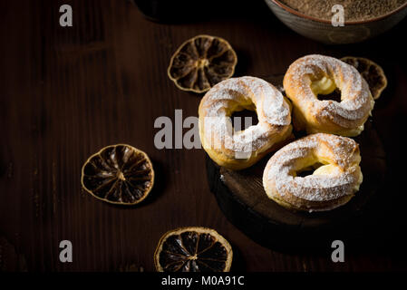 Horizontal photo of three cream puffs filled by cream and covered by powder and cinnamon sugar. Few dry orange rings are placed around on dark brown v Stock Photo