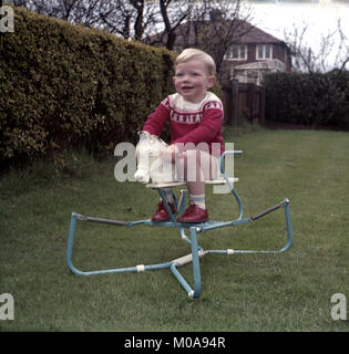 1960s, historical, small blond haired boy playing or bouncing on a metal spring loaded rocking horse sat on a lawn in a suburban back garden, England, UK. Stock Photo