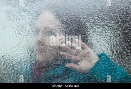 Woman looking through a window covered with thick, textured ice, with her hand on the window Stock Photo