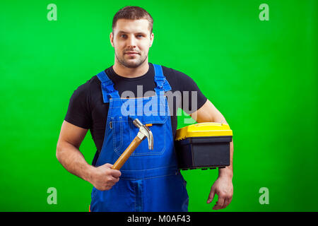 A dark-haired male construction worker in a black T-shirt and blue construction overall holds a wooden hammer and a box with construction tools on a g Stock Photo
