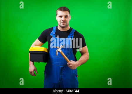A dark-haired male construction worker in a black T-shirt and blue construction overall holds a wooden hammer and a box with construction tools on a g Stock Photo