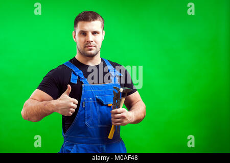 The dark-haired man builder in a black T-shirt and blue construction jumpsuit smiles while his thumb is up, and the other hand holds a wooden hammer a Stock Photo