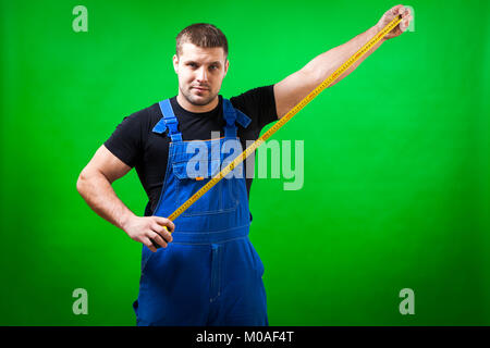 A dark-haired male builder in a black T-shirt and blue construction jumpsuit shows a long yellow tape rule on a green isolated background Stock Photo