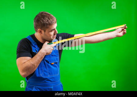 A dark-haired male builder in a black T-shirt and blue construction jumpsuit demonstrates and measures with his long yellow tape rule his hand on a gr Stock Photo