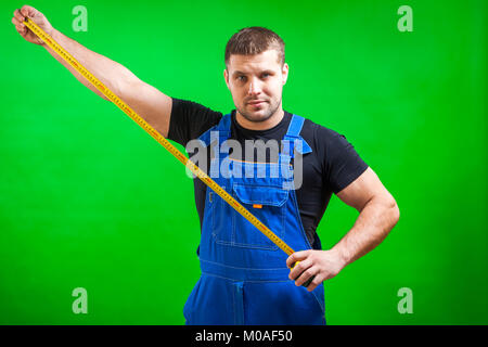 A dark-haired male builder in a black T-shirt and blue construction jumpsuit shows a long yellow tape rule on a green isolated background Stock Photo