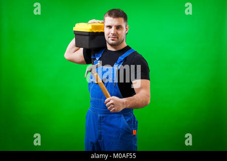 A strong man builder in a black T-shirt and blue construction jumpsuit holds a wooden hammer and a box with construction tools on his shoulder on a gr Stock Photo