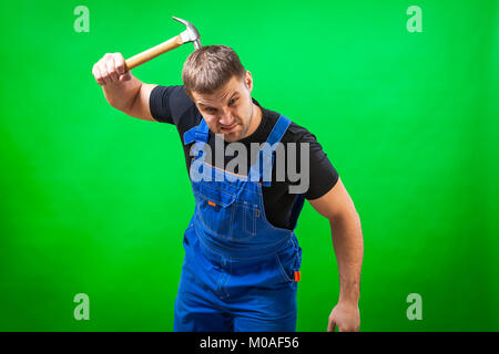 A cheerful male builder in a black T-shirt and blue construction jumpsuit laughs and scratches his head with a wooden hammer on a green isolated backg Stock Photo