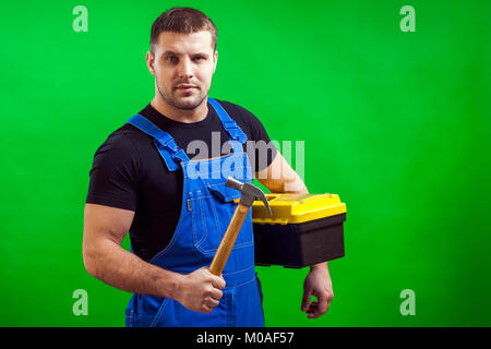 A dark-haired male construction worker in a black T-shirt and blue construction overall holds a wooden hammer and a box with construction tools on a g Stock Photo