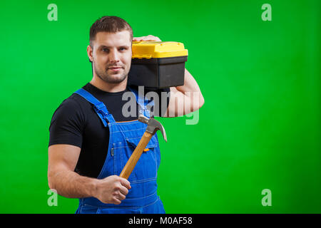 A strong man builder in a black T-shirt and blue construction jumpsuit holds a wooden hammer and a box with construction tools on his shoulder on a gr Stock Photo