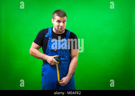 A dark-haired male construction worker wearing a black T-shirt and blue construction overall having fun, laughing funny  and uses  tape rule on a gree Stock Photo