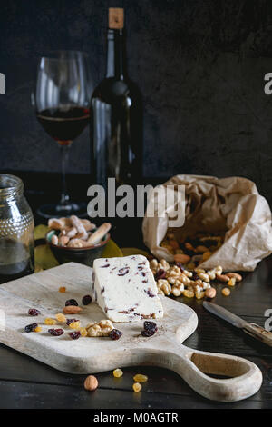 Wensleydale cheese with cranberries, red wine, honey, nuts, raisins on wooden cutting board. Black concrete background. Selective focus. Stock Photo