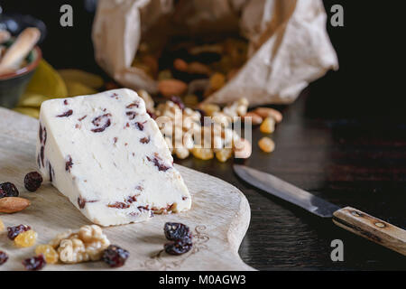 Wensleydale cheese with cranberries, red wine, honey, nuts, raisins on wooden cutting board. Black concrete background. Selective focus. Stock Photo