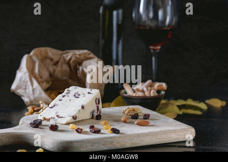 Wensleydale cheese with cranberries, red wine, honey, nuts, raisins on wooden cutting board. Black concrete background. Selective focus. Stock Photo