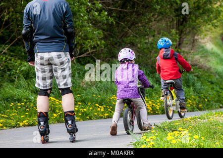 Children under parental supervision two toddlers ride away bikes followed father on roller skates Stock Photo