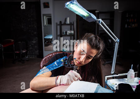 Master tattoo draws on the clients tattoo. Tattoo artist holding a metal tattoo machine in  sterile gloves and working on the professional blue mat. Stock Photo