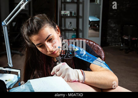 Master tattoo draws on the clients tattoo. Tattoo artist holding a metal tattoo machine in  sterile gloves and working on the professional blue mat. Stock Photo