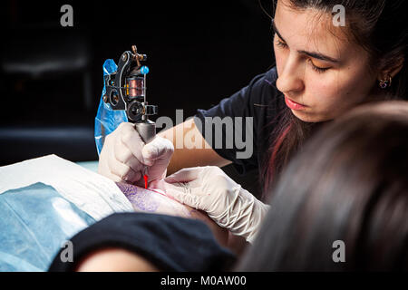 Master tattoo draws the red paint on the clients tattoo. Tattoo artist holding a metal tattoo machine in  sterile gloves and working on the profession Stock Photo