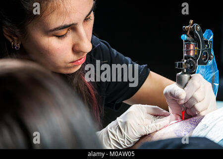 Master tattoo draws the red paint on the clients tattoo. Tattoo artist holding a metal tattoo machine in  sterile gloves and working on the profession Stock Photo