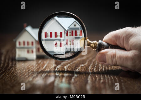 Hand Holding Magnifying Glass Up To Model Home on Reflective Wooden Surface. Stock Photo