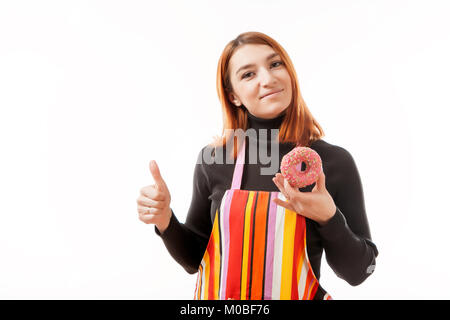 Young red-haired woman in black sweater and kitchen apron holds a pink donut with chocolate filling in her hand, shows it to the camera and smiles on  Stock Photo