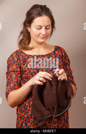 Close-up of a dark-haired pregnant woman in a summer dress in a floral print smiling and knitting with knitting needles from a natural sherry thread a Stock Photo