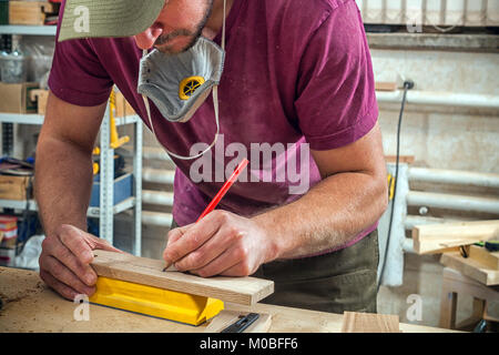 A young strong man builder carpenter works with a wooden bar for making furniture, measures and cuts in the workshop, in the background of many tools  Stock Photo