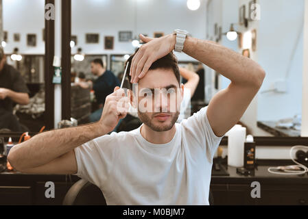 Working process in modern barbershop. Handsome hairdresser serving customer, styling hair for male client using hairdryer. Side view portrait of attra Stock Photo