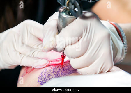 Close-up of a young woman tattoo master in sterile gloves makes a tattoo on the thigh with red paints Stock Photo